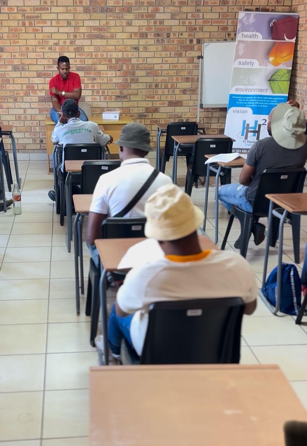 Group of people sitting in classroom desks with a brick wall and educational posters in the background.